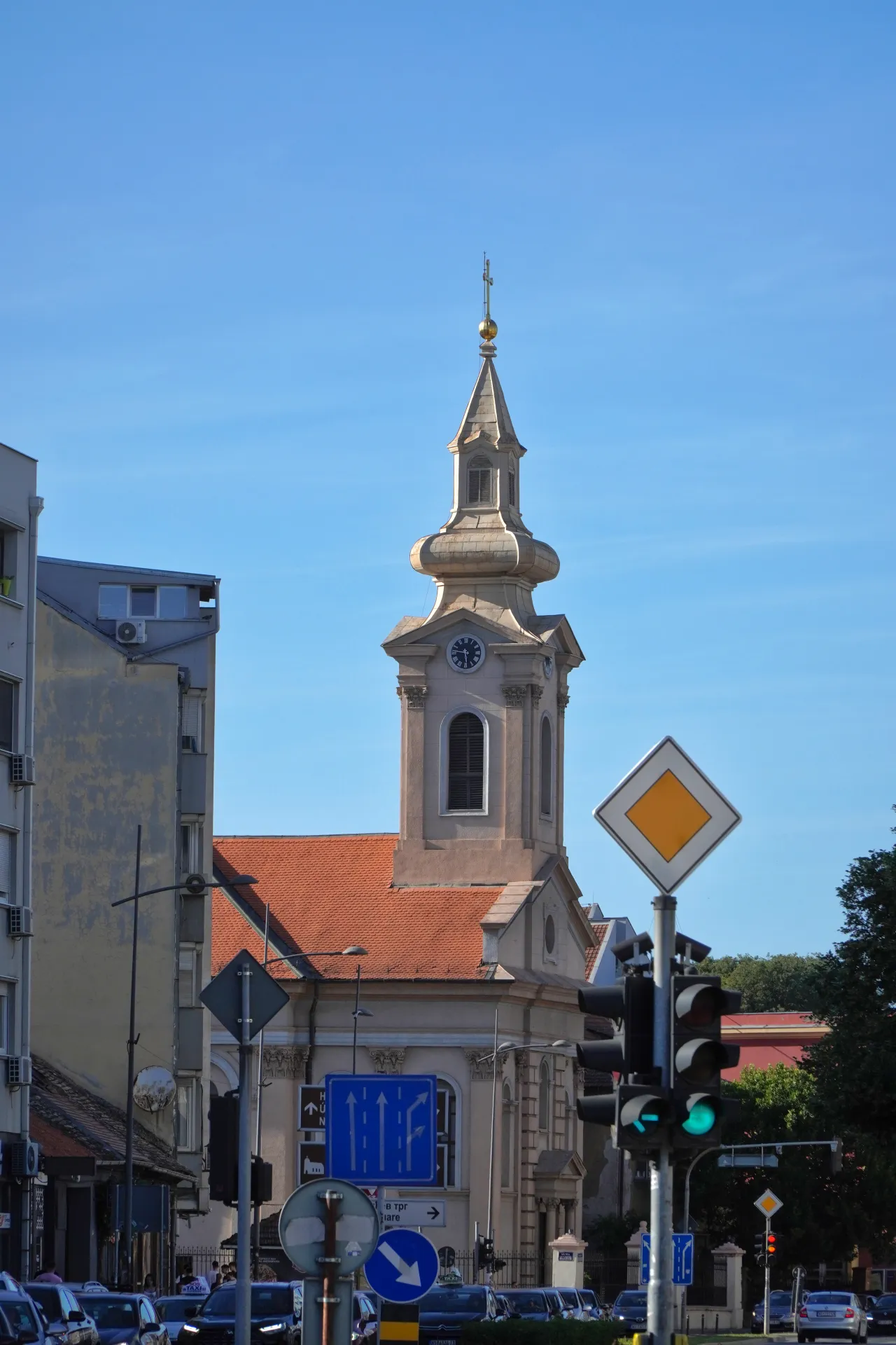 A bell tower of a church with a clock and a gold cross on top, surrounded by buildings, traffic lights, and road signs on a sunny day in Novi Sad, Serbia.