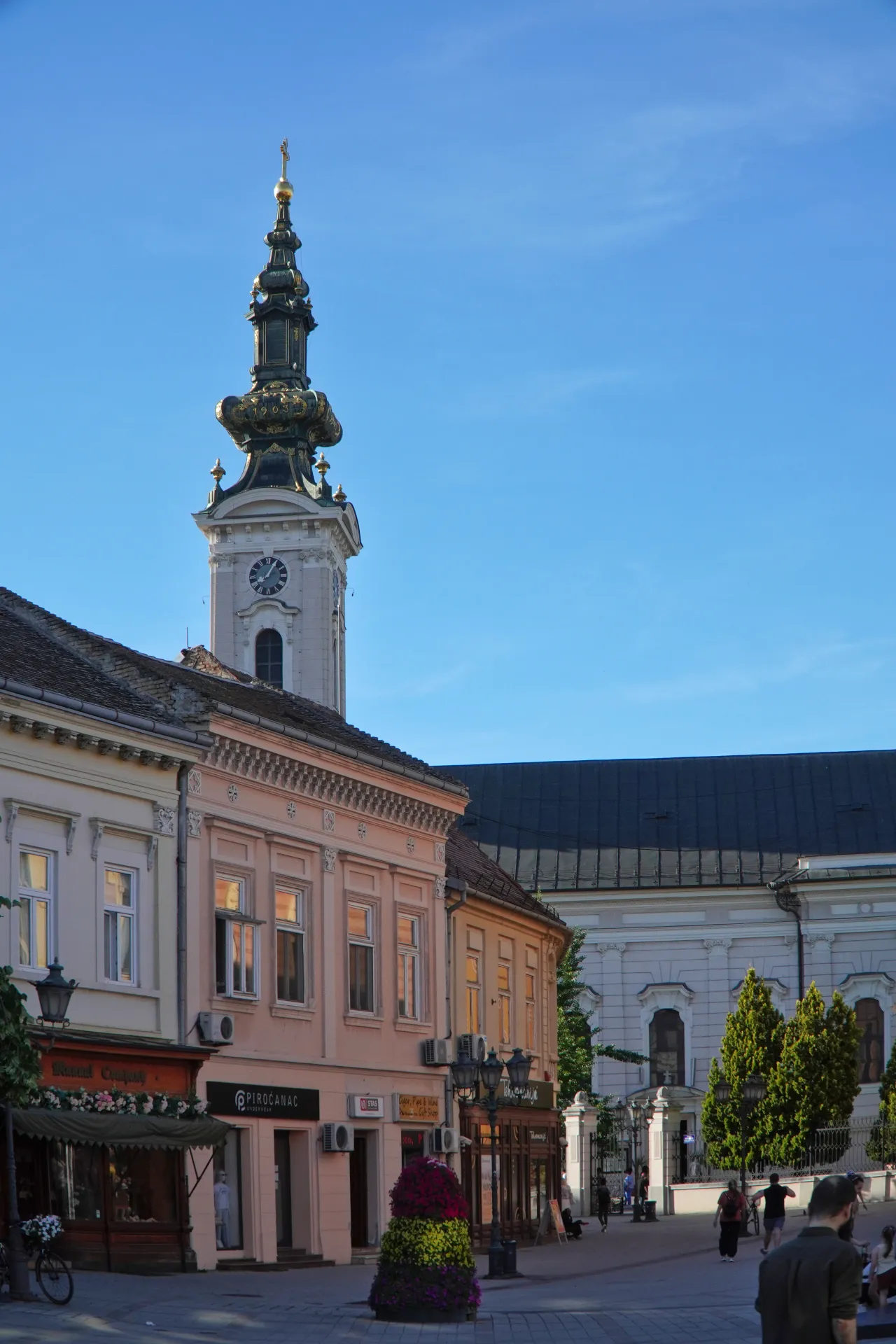 A sunny street in Novi Sad, Serbia, with colorful buildings, a tall church clock tower, and people walking by.