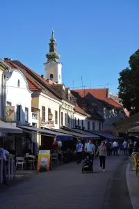 People walking on a cobblestone street in Zagreb, with traditional buildings and a prominent church tower in the background.