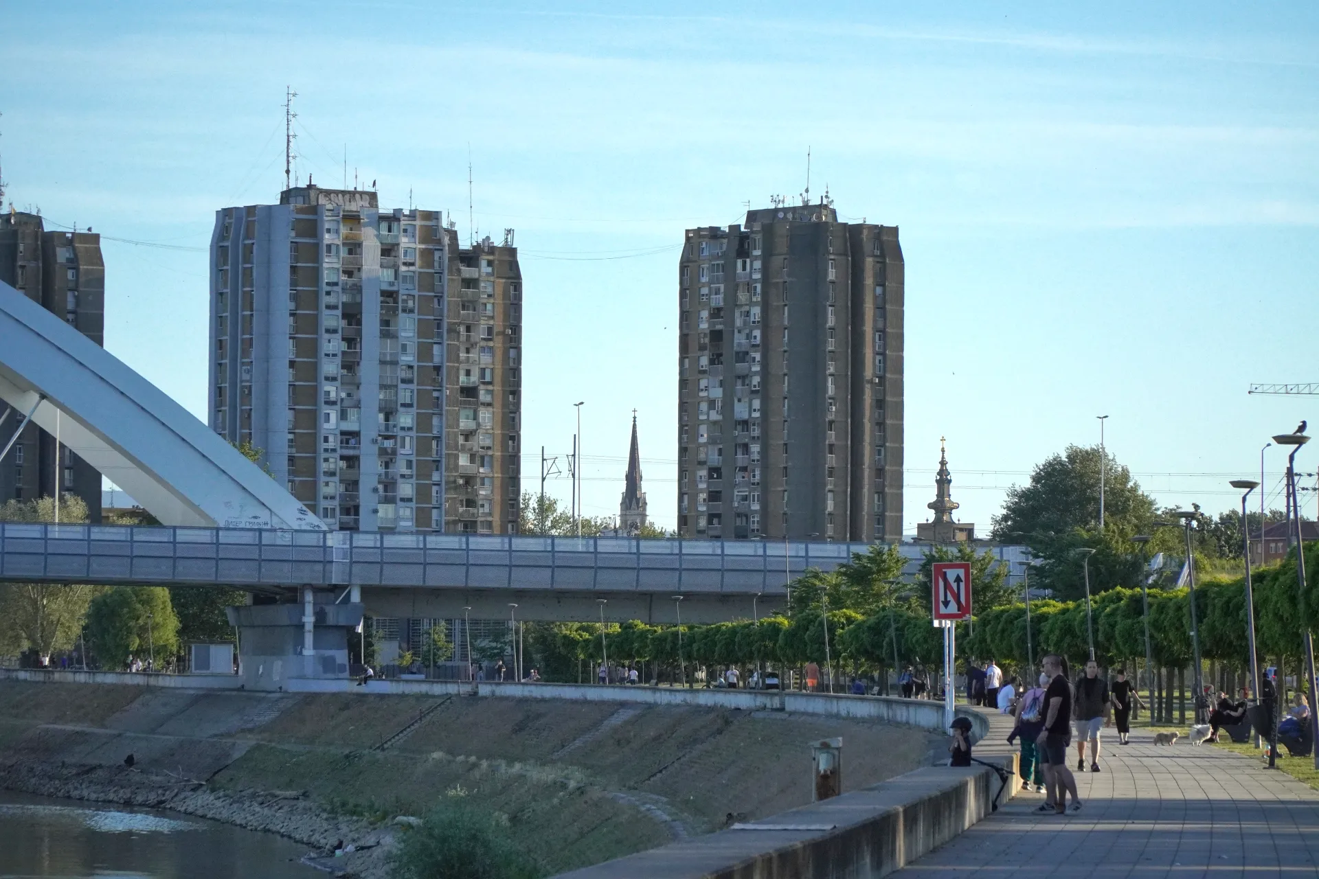 Tall concrete apartment buildings stand against a clear blue sky in Novi Sad, Serbia, with a large white bridge spanning across the foreground. People are seen walking along the paved riverfront promenade, bordered by green trees and lampposts.
