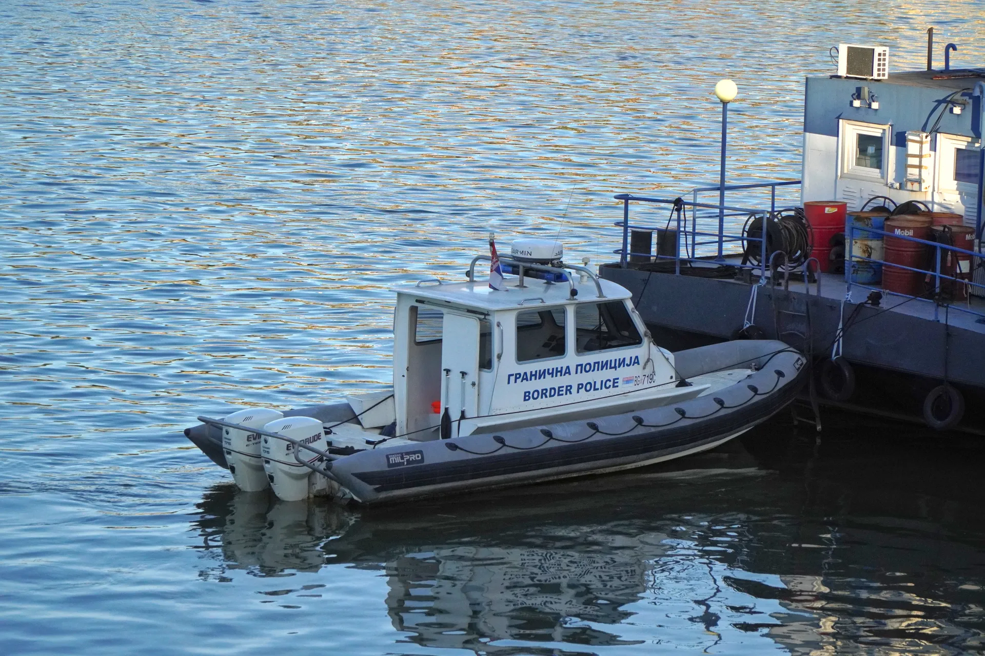 Бело-серая надувная лодка с надписью 'BORDER POLICE' и 'ГРАНИЧНА ПОЛИЦИЈА' кириллицей, а также сербским флагом, привязана к большему темному катеру на реке Дунай в Нови-Саде, Сербия. Вода спокойная, с отражениями.