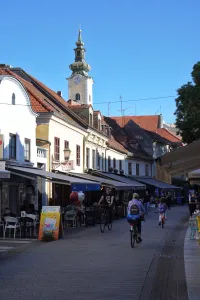 A sunny street in Zagreb, Croatia, with people cycling past cafes, and the clock tower of St. Mark's Church in the background.