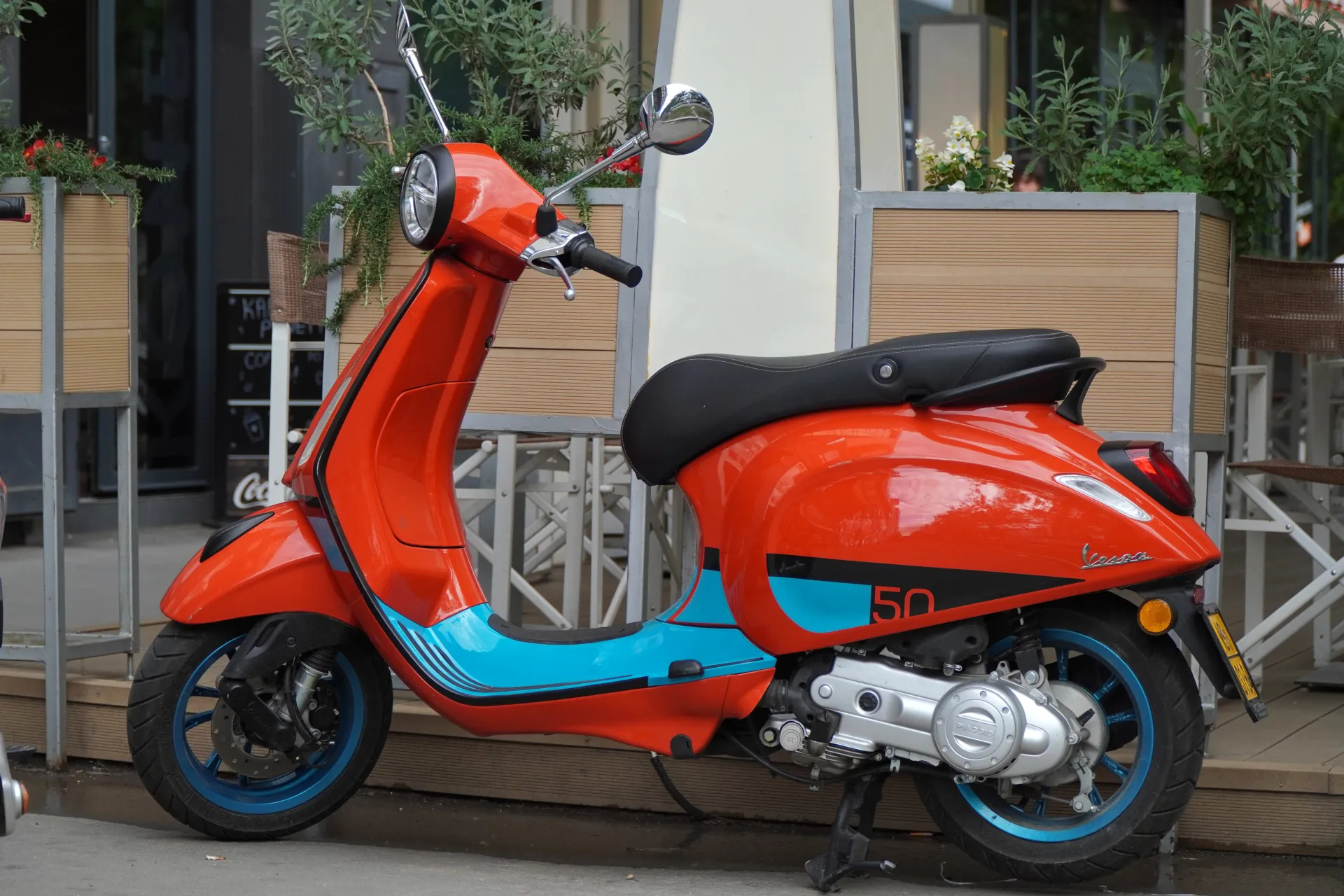 An orange Vespa scooter with blue trim and a black seat is parked on a wet sidewalk in front of a building in Novi Sad, Serbia, with green plants and a cafe in the background.