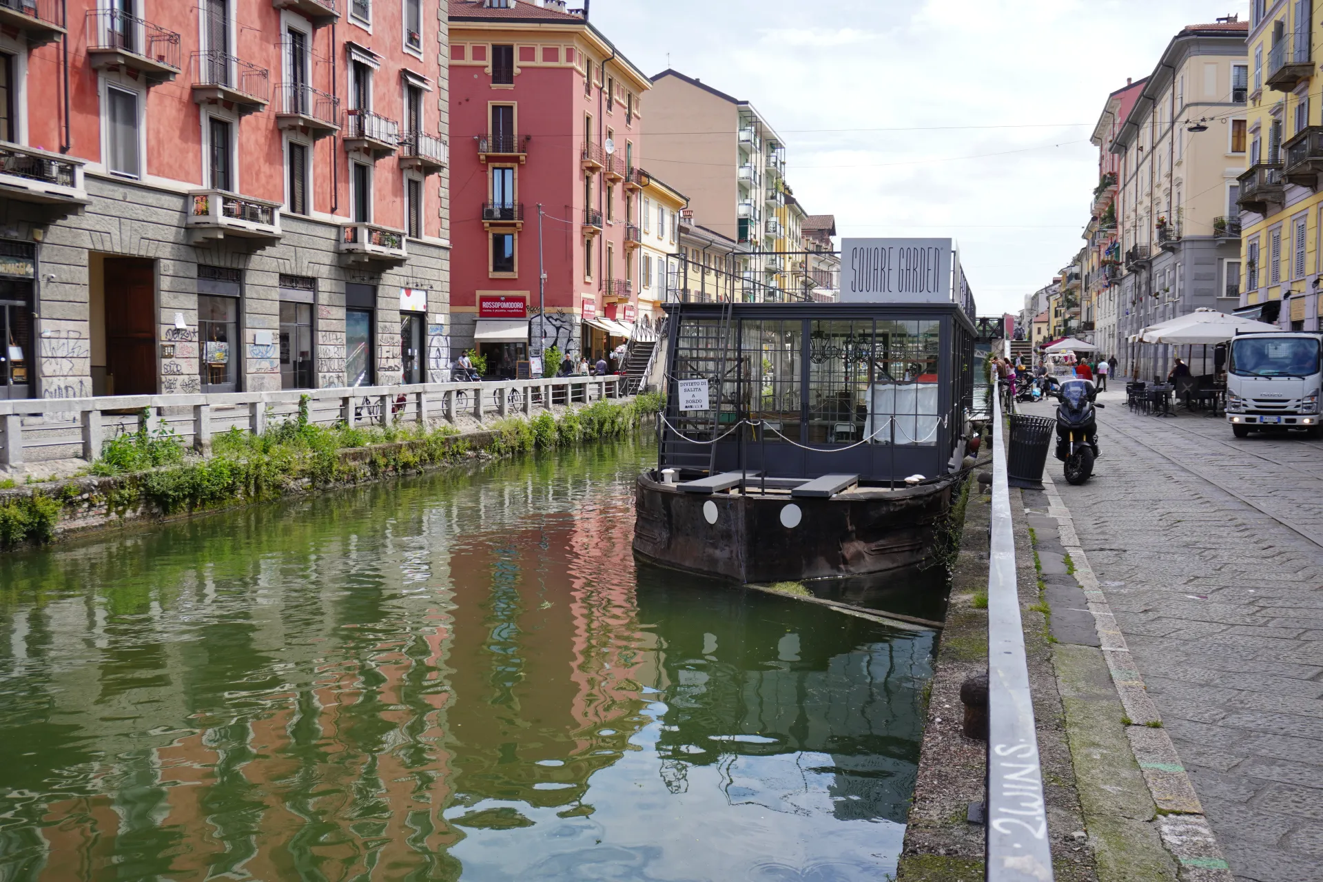 Kanal u milanskom distriktu Navigli sa šarenim zgradama na levoj strani, brodom sa natpisom 'Square Garden', skuterom i kamionom na desnoj strani popločane ulice.