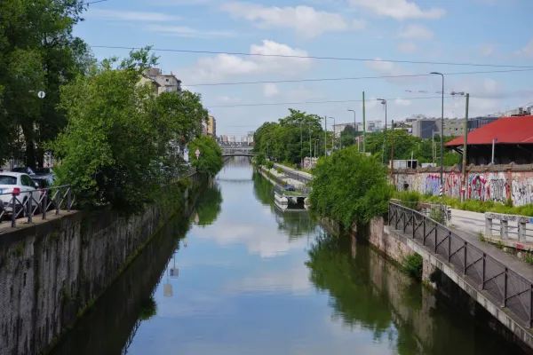 Un canal ancho en Milán, Italia, con árboles verdes bordeando las orillas, muros de piedra y edificios modernos al fondo. El cielo y los árboles se reflejan en el agua quieta, y un puente cruza el canal a lo lejos.
