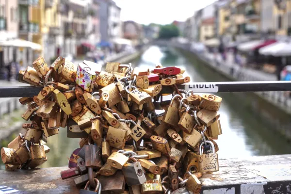 Una pila de candados desgastados y coloridos unidos a cadenas en la barandilla de un puente, con un canal y edificios de Milán desenfocados al fondo.
