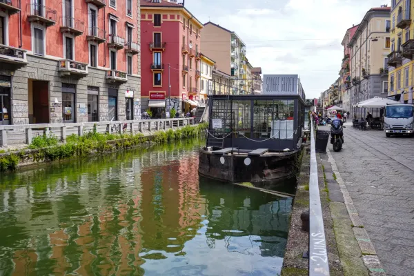 Un canal en el distrito de Navigli de Milán con edificios coloridos a ambos lados, una pasarela peatonal, gente caminando, un bote en el agua y un puente al fondo.