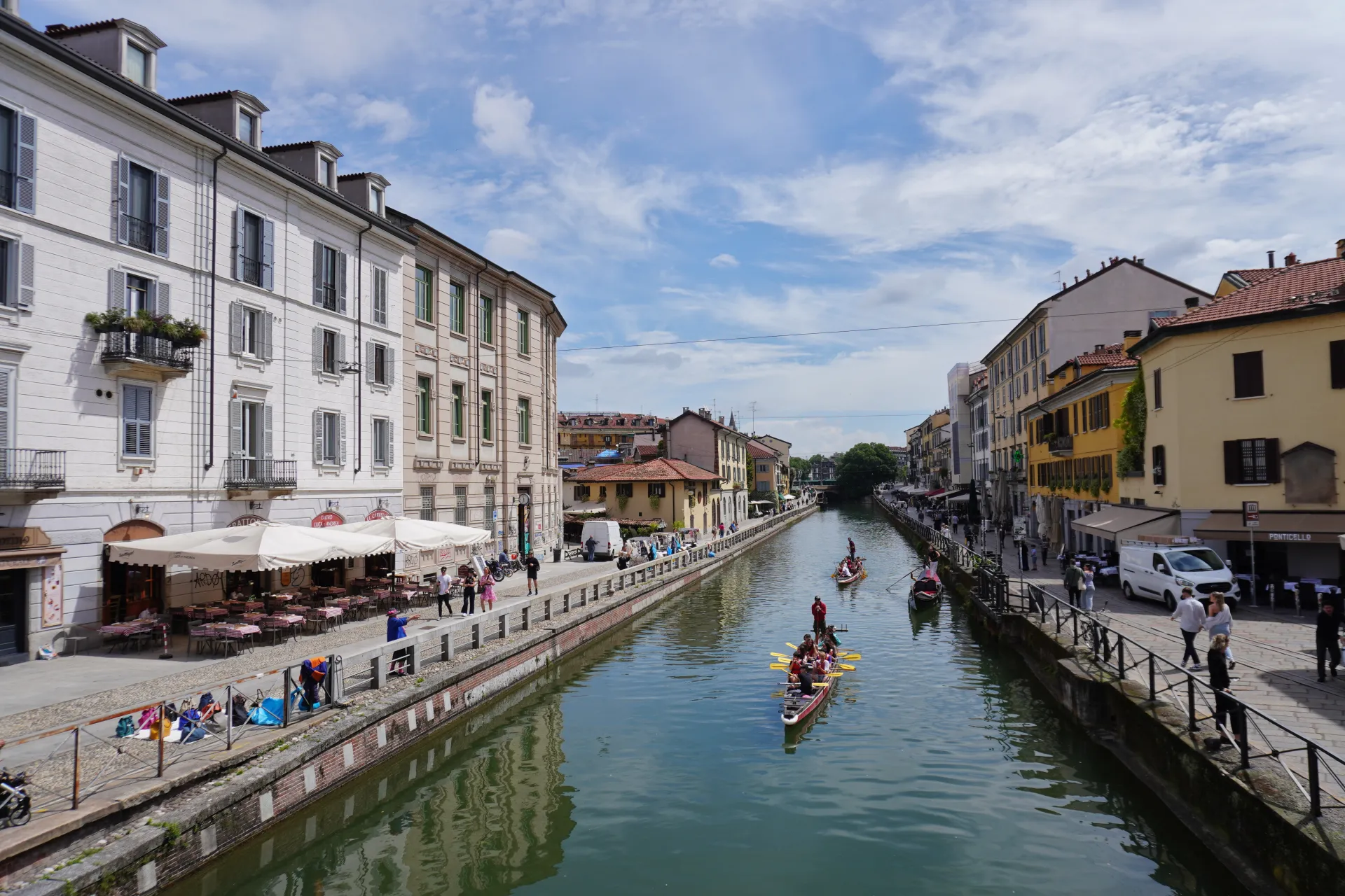 People rowing boats down a canal lined with historic buildings and outdoor cafes in Milan's Navigli district.