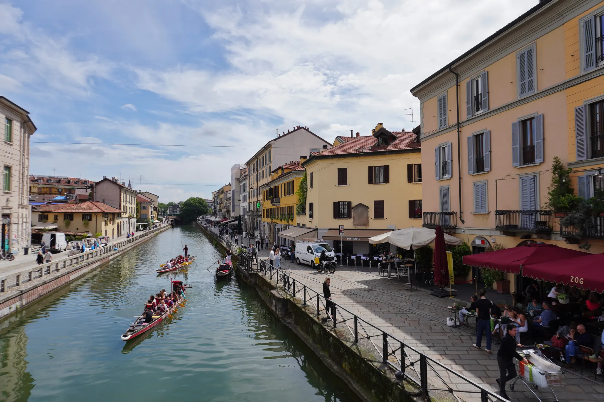 A canal in Milan's Navigli district with dragon boats racing, flanked by colorful buildings and outdoor seating areas.