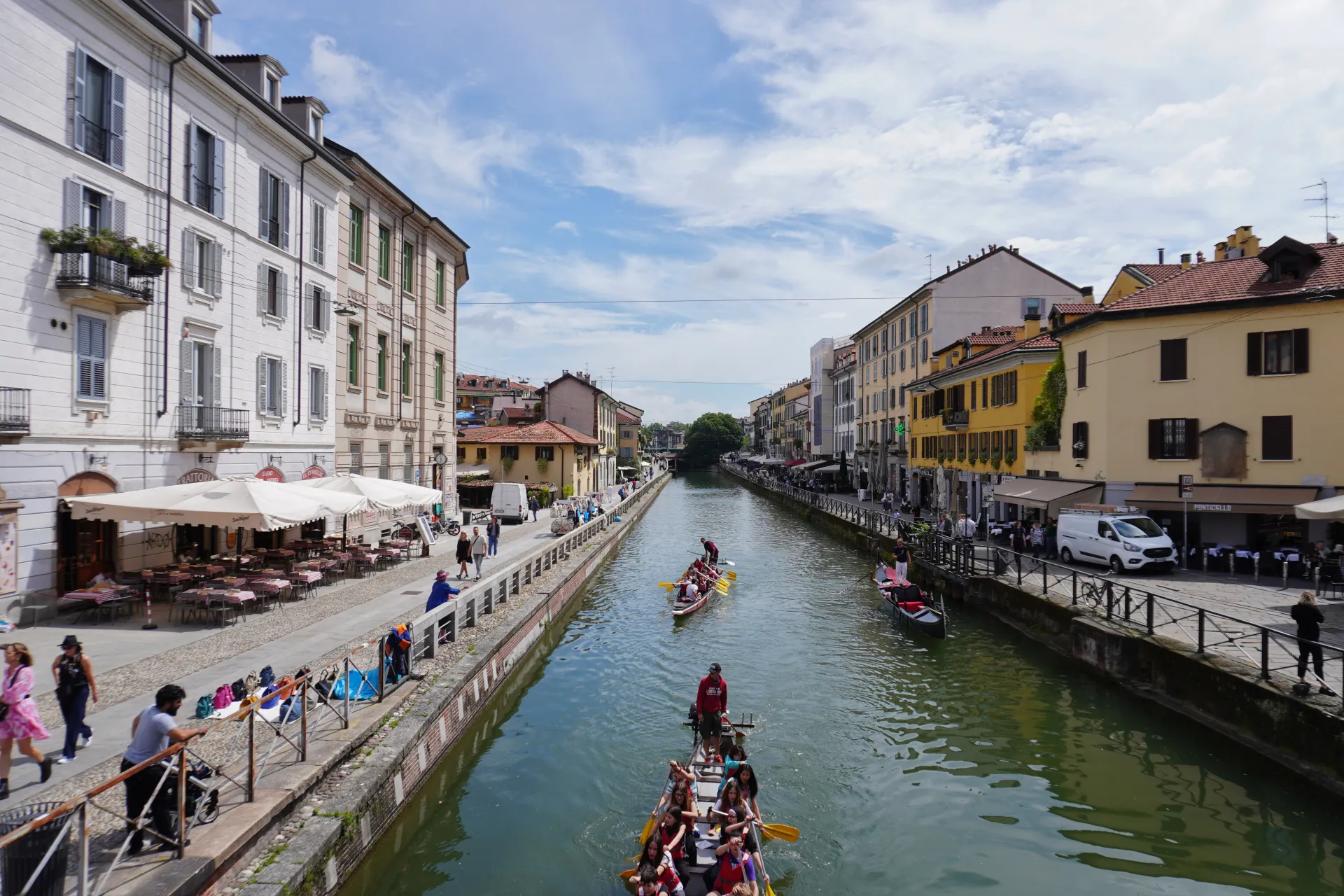 A sunny day in Milan's Navigli district, showing a canal lined with buildings and people on boats.