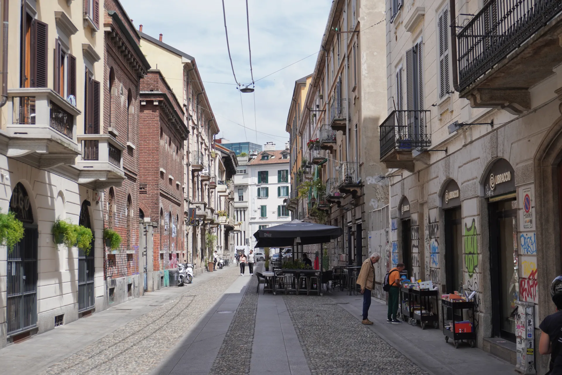 A view down a cobblestone street in Milan, Italy, flanked by colorful buildings with wrought-iron balconies and closed shutters.
