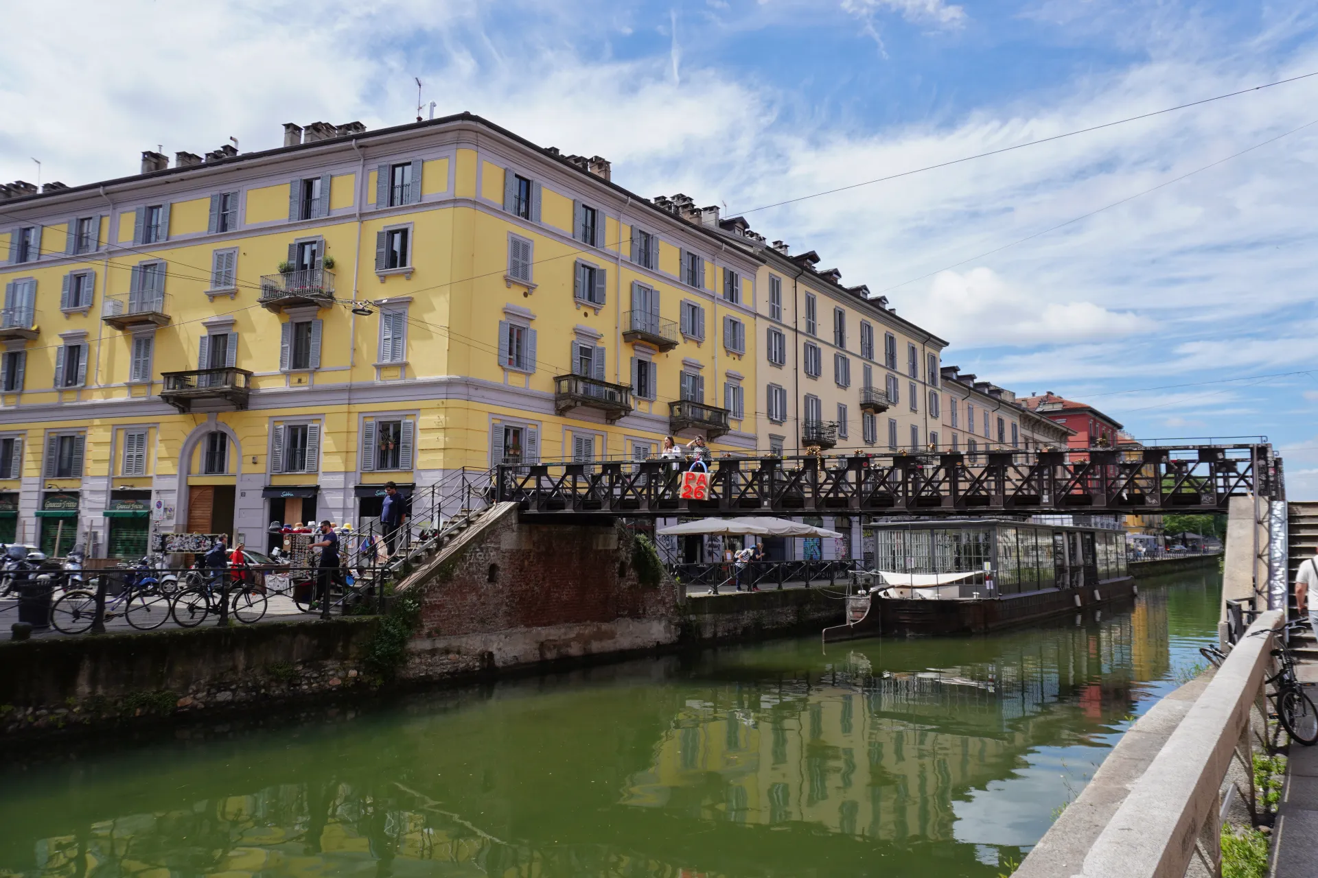 A wide canal in Milan's Navigli district lined with yellow and beige buildings, with a pedestrian bridge crossing over it. Boats are docked on the canal, and people are visible on the bridge and along the canal path.