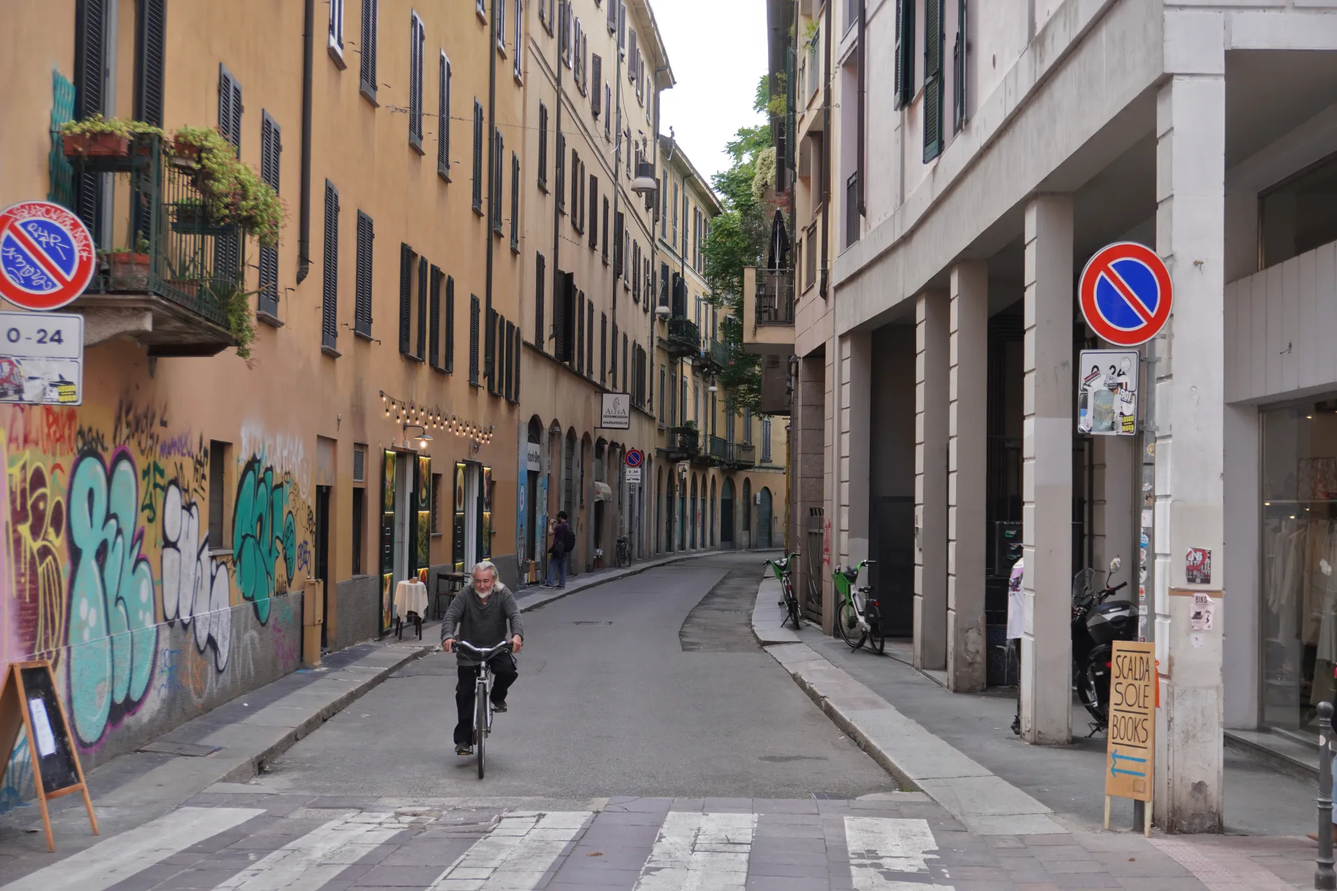 A man on a bicycle crosses a pedestrian street in Milan, Italy, with buildings on both sides showing shutters, balconies, graffiti, and shops.