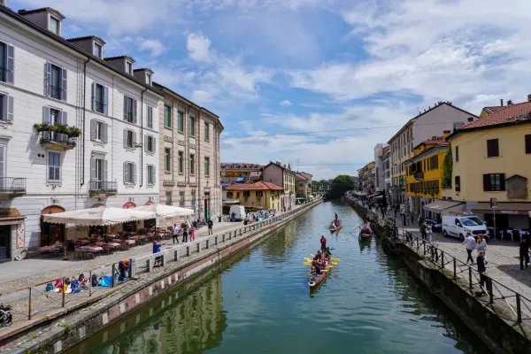 Un amplio canal en el distrito de Navigli de Milán, bordeado por edificios amarillos y beige, con un puente peatonal que lo cruza. Hay barcos atracados en el canal y personas visibles en el puente y a lo largo del camino del canal.
