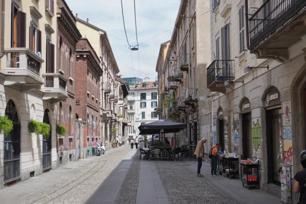 Ein Mann auf einem Fahrrad überquert eine Fußgängerstraße in Mailand, Italien. Gebäude mit Fensterläden, Balkonen, Graffiti und Geschäften säumen die Gasse.