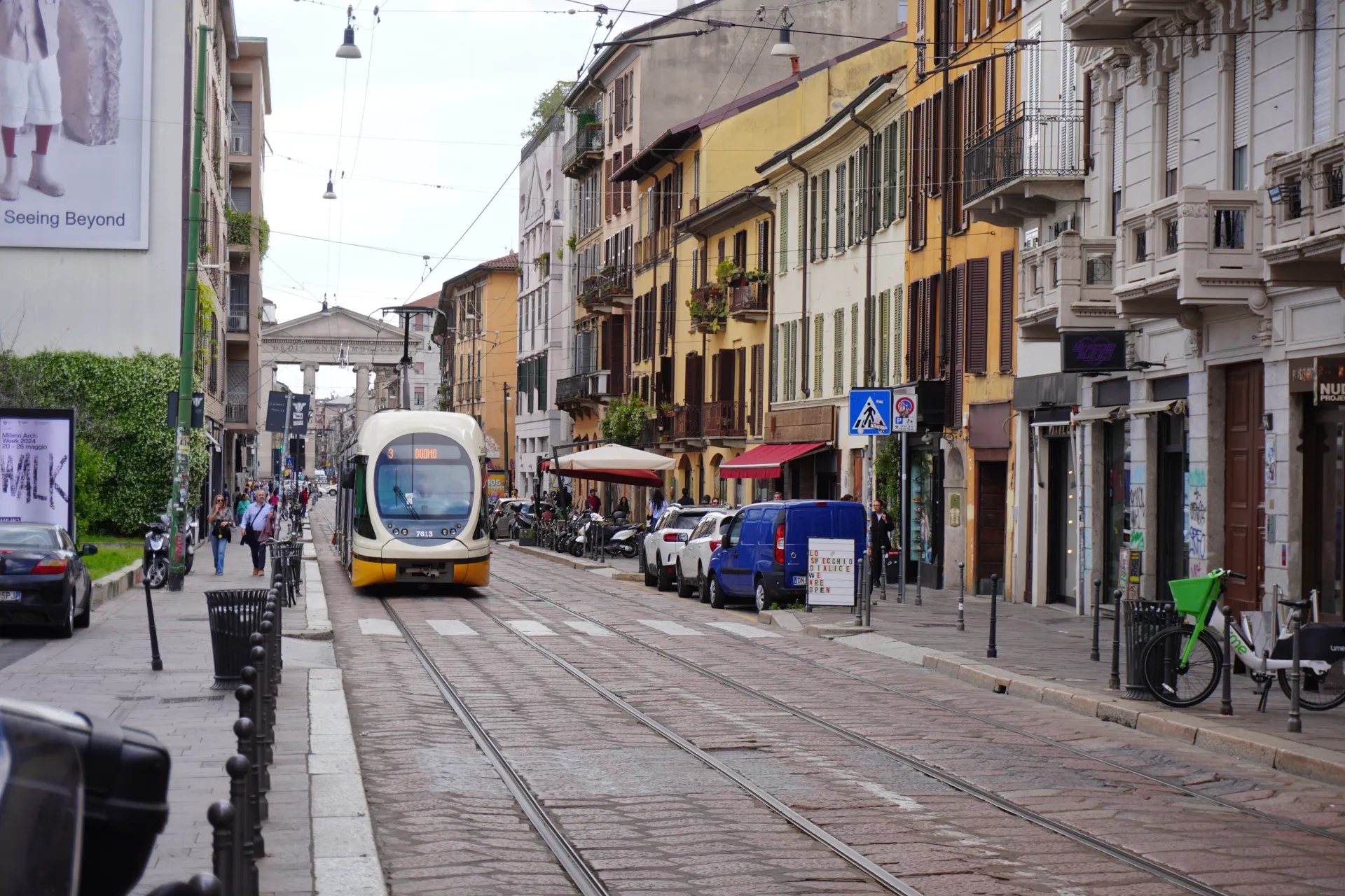 A yellow and white tram travels down a street in Milan with vintage buildings and shops lining the sides.