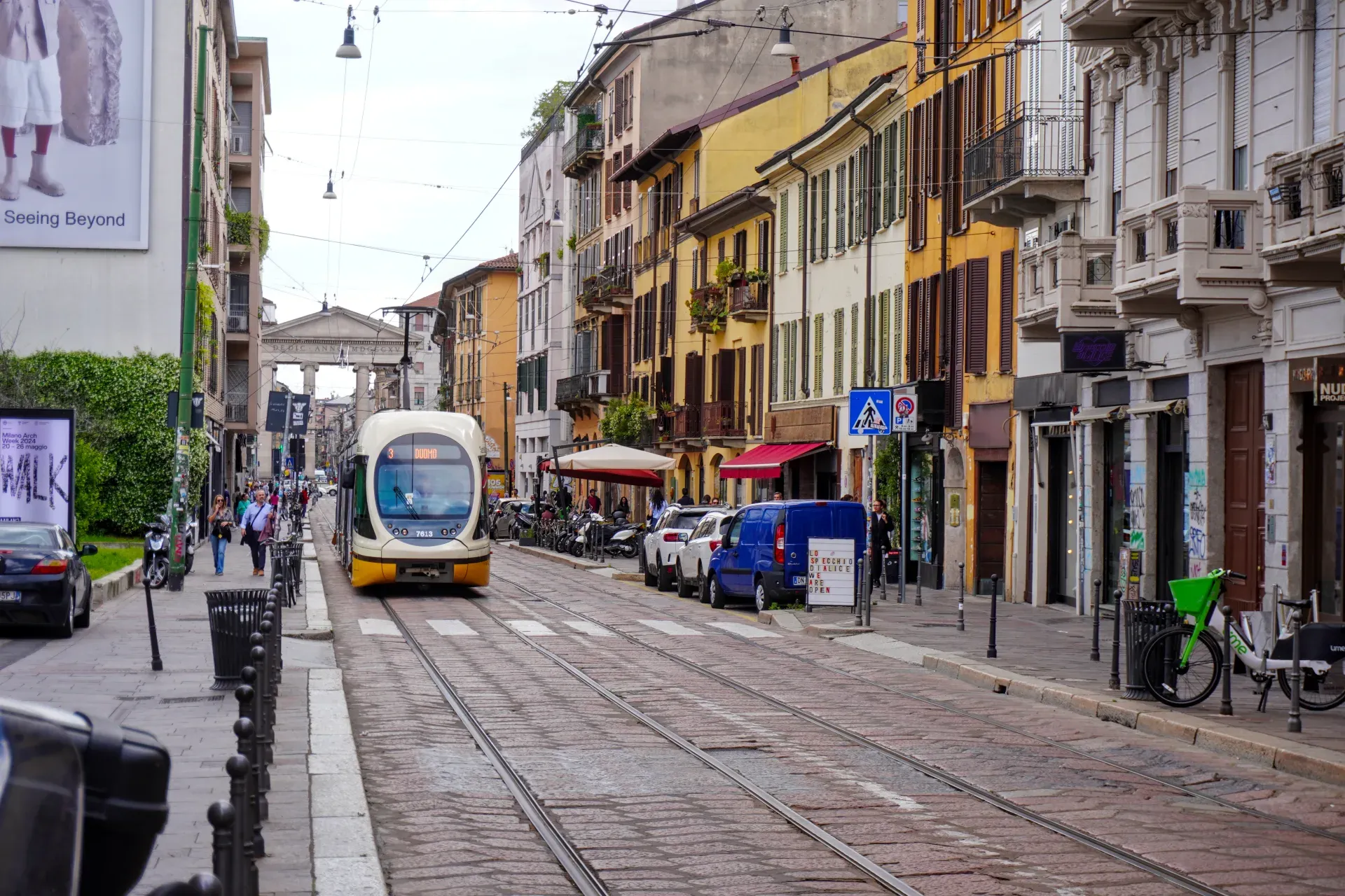 Žuti i beli tramvaj prolazi ulicom u Milanu, sa starinskim zgradama i prodavnicama sa strane.
