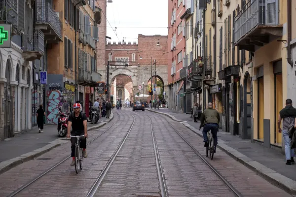 Un tranvía verde con la marca 'Foxtown' circula por una calle de Milán, Italia, con peatones en la acera y un ciclista cerca. Edificios históricos y un arco son visibles al fondo.