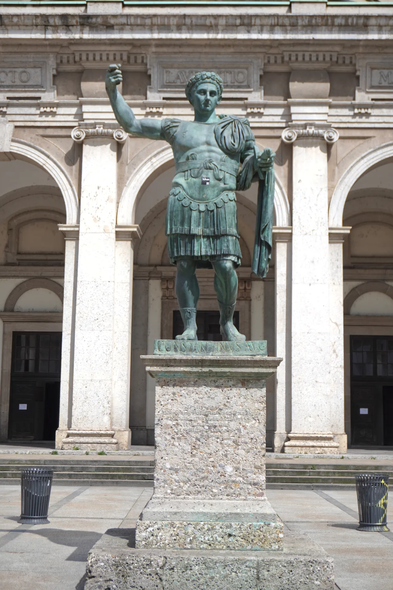 Visoka bronzana statua cara Konstantina Velikog u vojnom oklopu, koja stoji na kamenom postolju ispred zgrade sa grandioznim lukovima i klasičnom arhitekturom u Milanu, Italija.