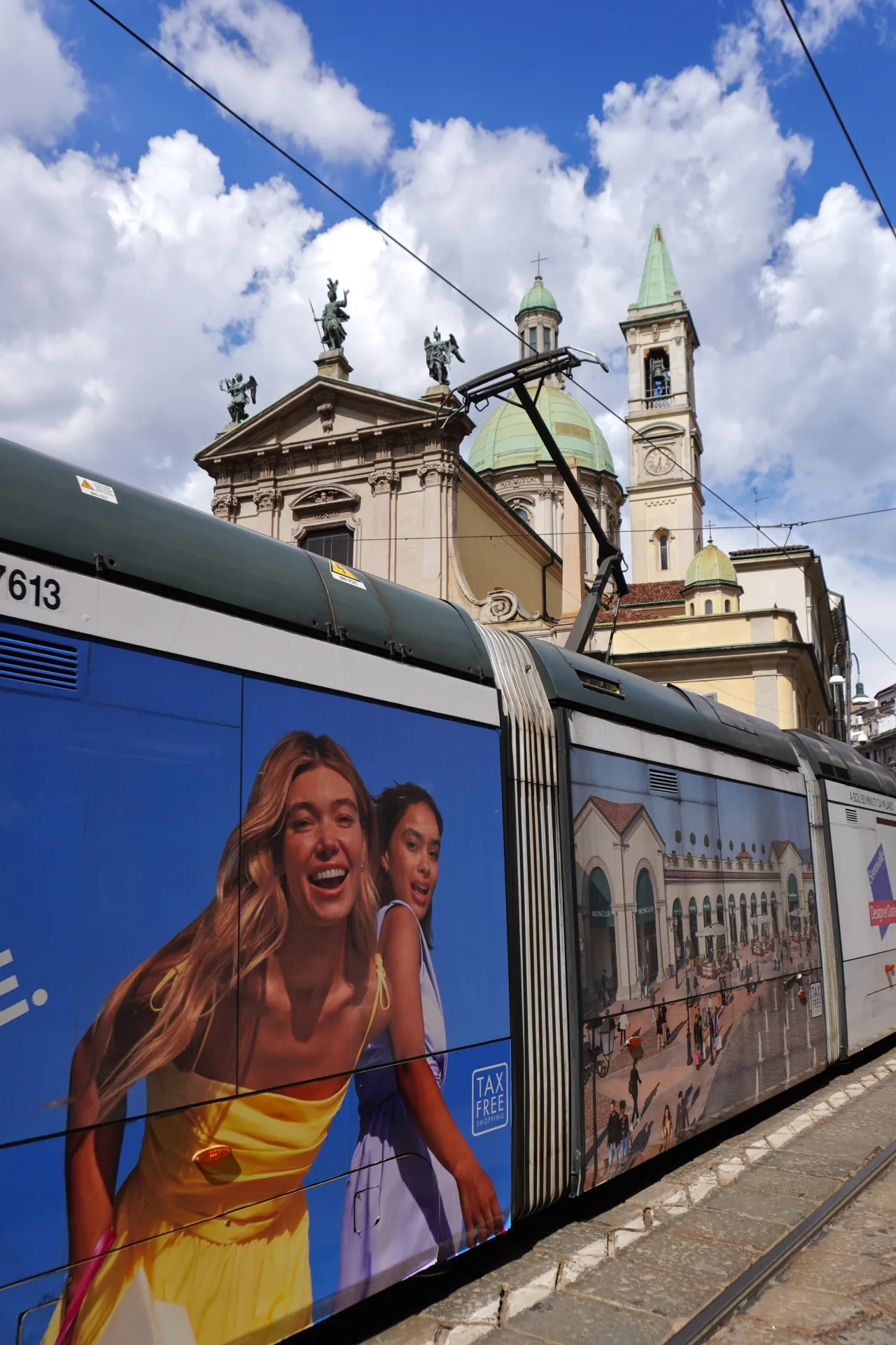 A tram in Milan, Italy, with colorful advertisements on its side, passing by a church and historical buildings under a bright, cloudy sky.