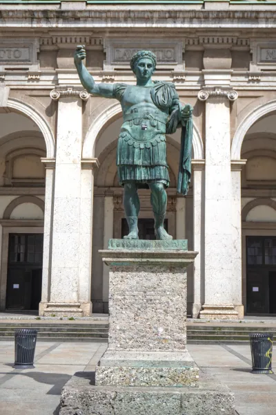 Una alta estatua de bronce del emperador Constantino el Grande con armadura militar, de pie sobre un pedestal de piedra frente a un edificio con grandes arcos y arquitectura clásica en Milán, Italia.