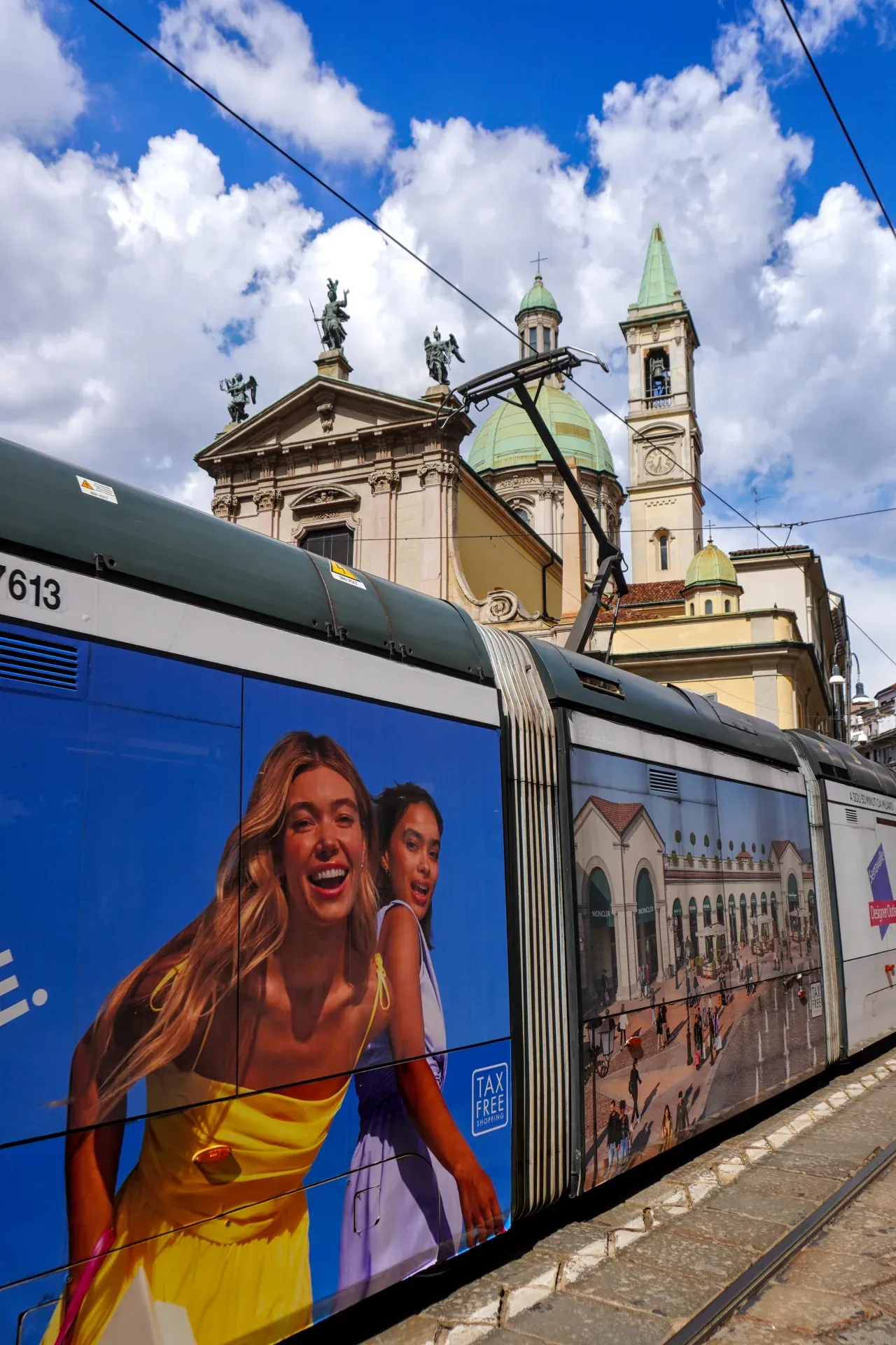 A tram in Milan, Italy, adorned with colorful advertisements on its sides, passes by a church and historic buildings under a bright, cloudy sky.