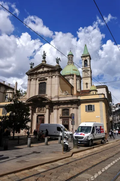 Una iglesia europea con cúpula y arquitectura clásica se alza en una calle de Milán, con vías de tranvía en primer plano y una furgoneta de reparto de FedEx estacionada bajo un cielo azul con nubes.