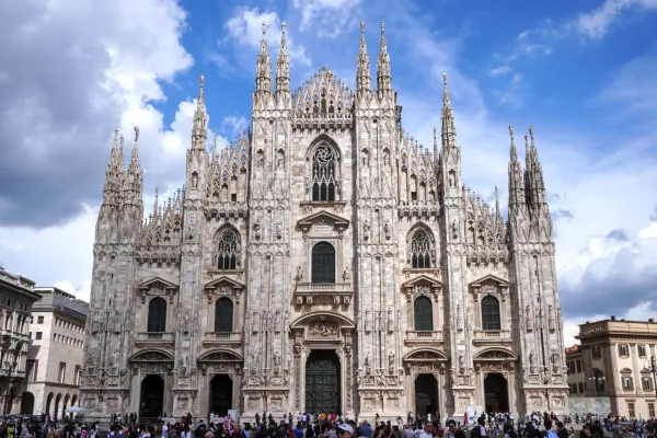 La ornamentada fachada de la Catedral de Milán, una gran iglesia gótica, con muchas agujas y estatuas, bajo un cielo azul con nubes. Una multitud de personas se reúne en la plaza frente a la catedral.