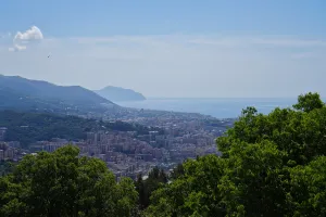 A wide aerial view of Genoa, Italy, showing buildings on green hills extending to the Ligurian Sea under a blue sky with a bird flying.