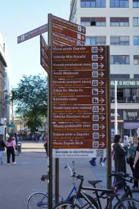 A brown street signpost in Zagreb, Croatia, with multiple directional signs in Croatian and English, pointing to various museums, galleries, and other points of interest, with bicycles parked in the foreground and a busy street scene in the background.