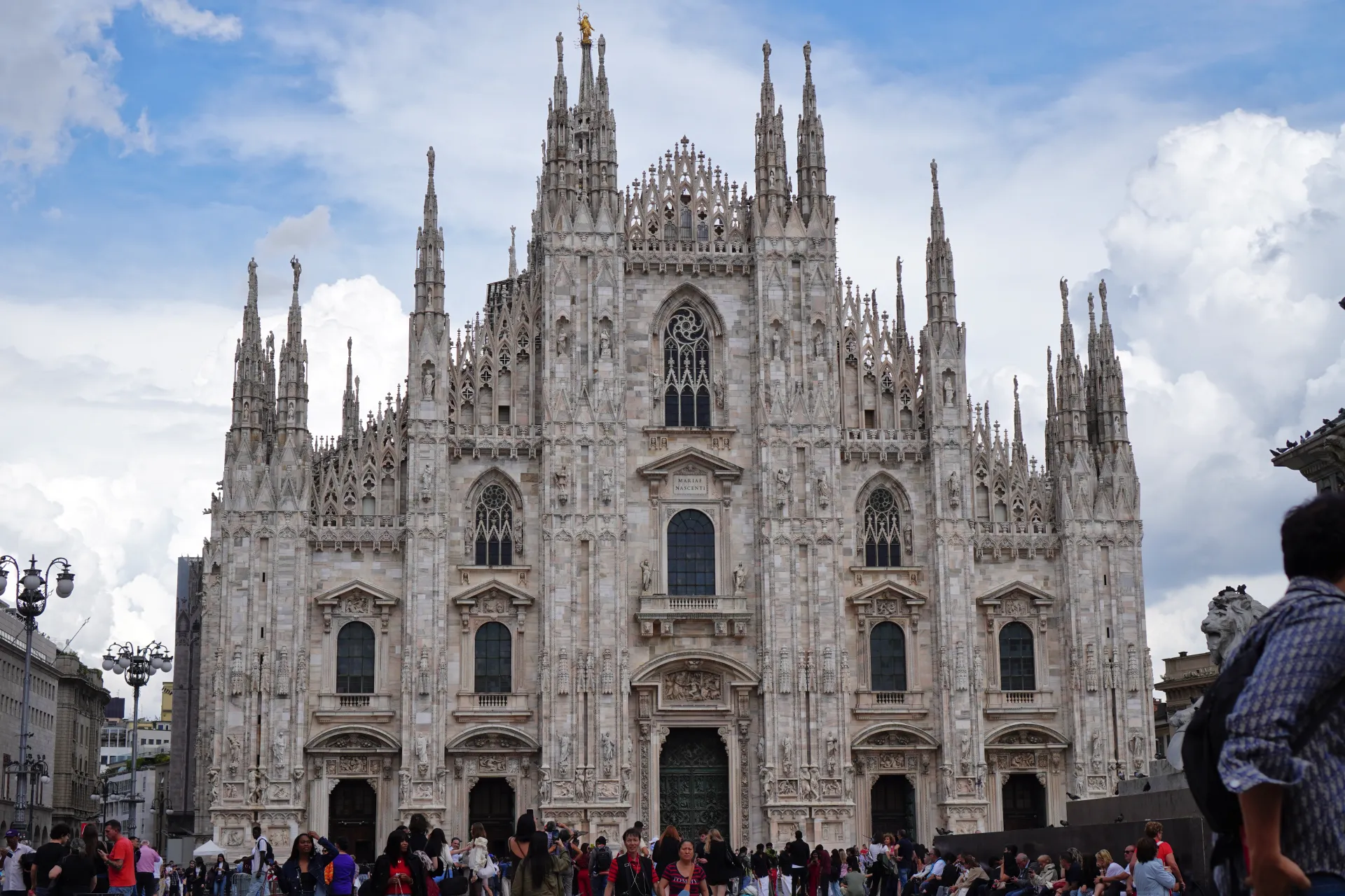Širok kadar Duomo di Milano, velike gotičke katedrale sa brojnim tornjevima i zamršenim kamenim rezbarijama, na pozadini oblačnog neba. Na prednjem planu na Piazza del Duomo vidljivi su ljudi.