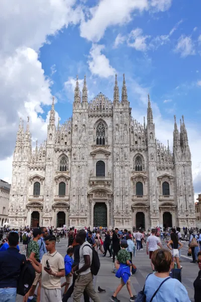La fachada ornamentada de la Catedral de Milán, Italia, con muchas agujas y detalles de piedra bajo un cielo azul brillante con nubes dispersas. Una multitud de personas es visible en primer plano en la plaza.