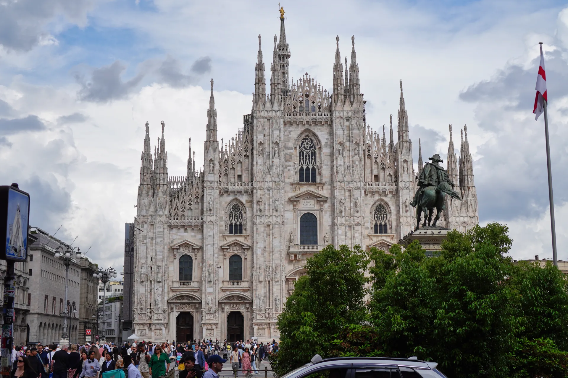 Slojevita fasada Milanske katedrale (Duomo di Milano) pod oblačnim nebom, sa bronzanom konjičkom statuuom i drvećem ispred, i gomilom ljudi na trgu.