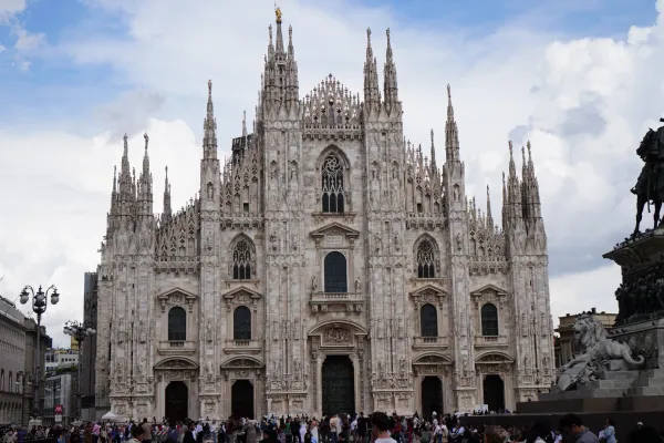 La intrincada fachada de la Catedral de Milán (Duomo di Milano) bajo un cielo nublado, con una estatua ecuestre de bronce y árboles delante, y una multitud en la plaza.
