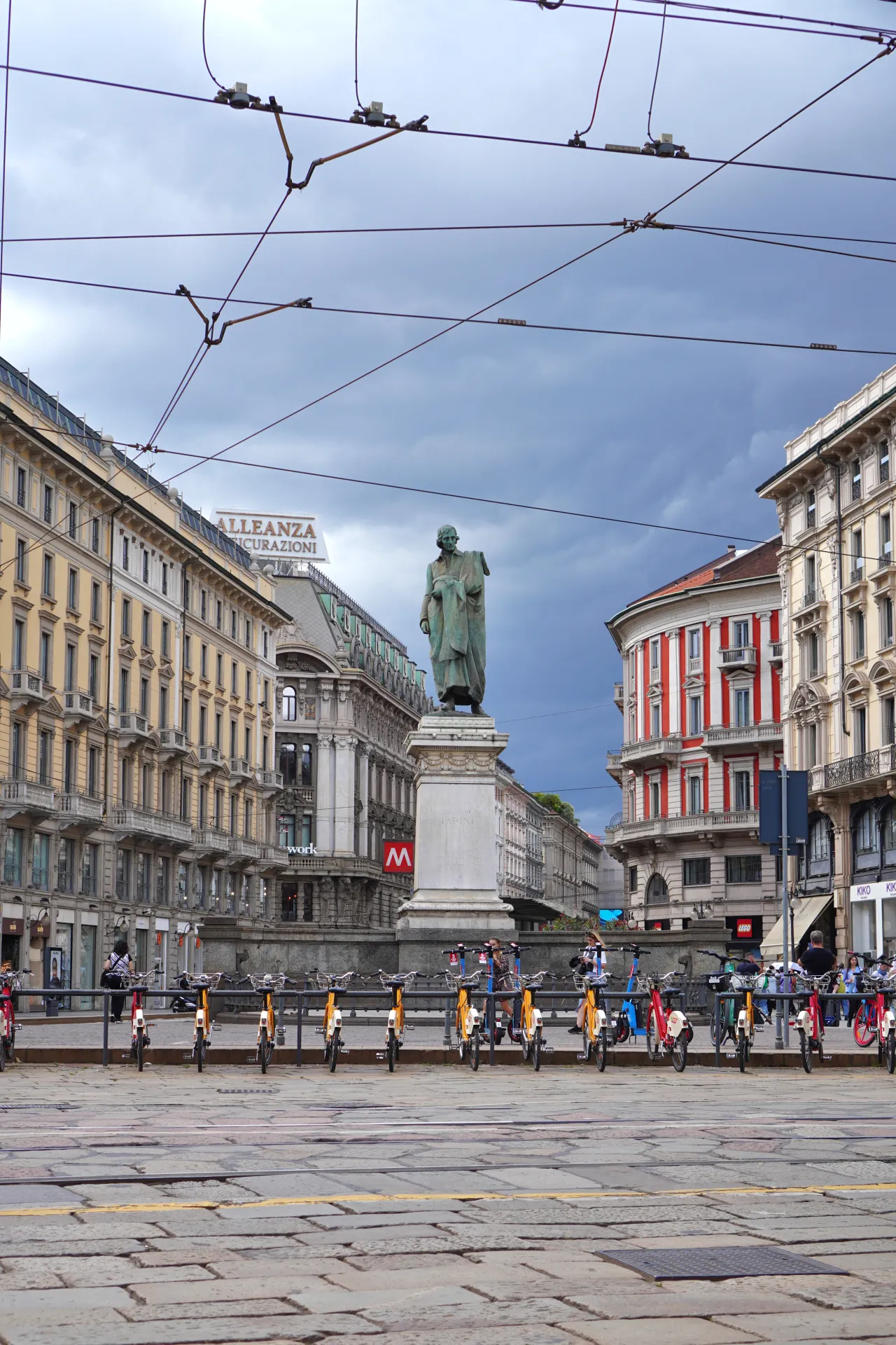 A wide shot of Piazza Cavour in Milan, Italy, dominated by a bronze statue on a pedestal, with ornate buildings lining the square and a line of shared bicycles in the foreground.