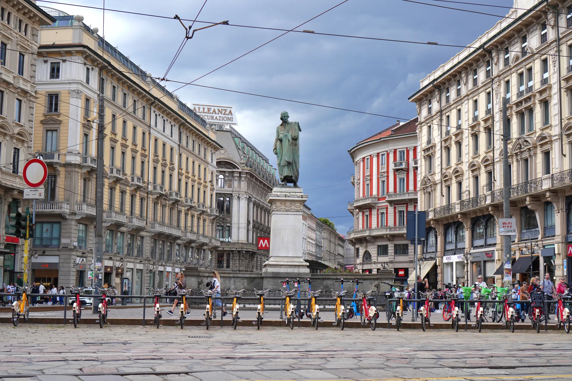 A street in Milan, Italy, with ornate buildings lining the sides, a bronze statue on a pedestal in the center, and numerous yellow and red electric scooters and bikes parked in a row.