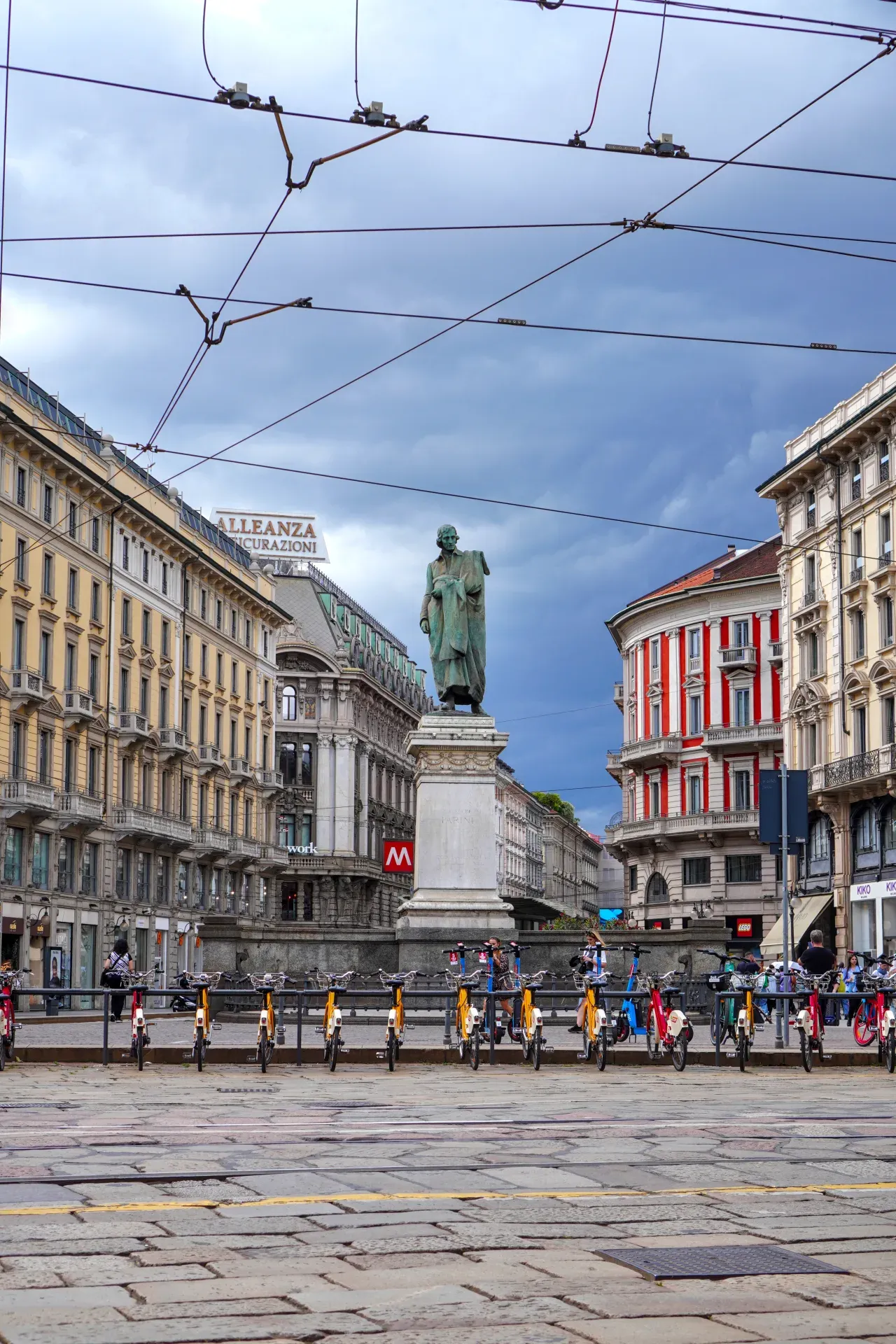 A wide shot of Piazza Cavour in Milan, Italy, dominated by a bronze statue on a pedestal, with ornate buildings lining the square and a row of rental bikes in the foreground.