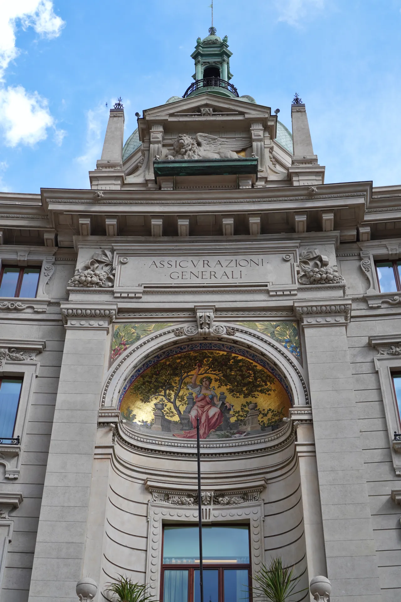 A detailed view of the upper facade of the Assicurazioni Generali building in Milan, with classical architectural elements, a mosaic, and a winged lion.