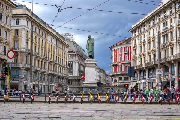 Eine Straße in Mailand, Italien, mit verzierten Gebäuden, die die Seiten säumen, einer Bronzestatue auf einem Sockel in der Mitte und zahlreichen gelben und roten Elektrorollern und Fahrrädern, die in einer Reihe geparkt sind.
