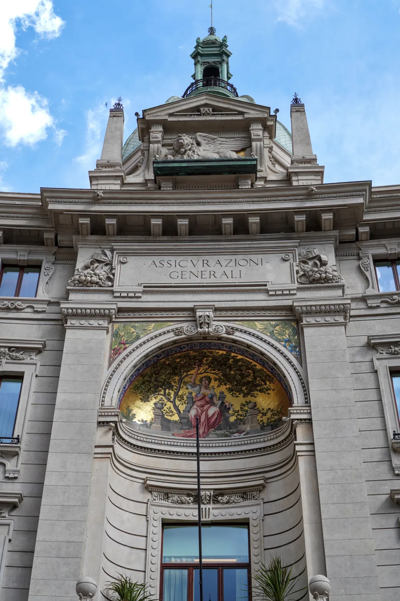 Une vue détaillée de la façade supérieure du bâtiment Assicurazioni Generali à Milan, présentant des éléments architecturaux classiques, une mosaïque et un lion ailé.