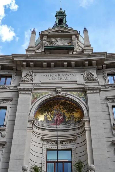 Una vista detallada de la fachada superior del edificio Assicurazioni Generali en Milán, que muestra elementos arquitectónicos clásicos, un mosaico y un león alado.