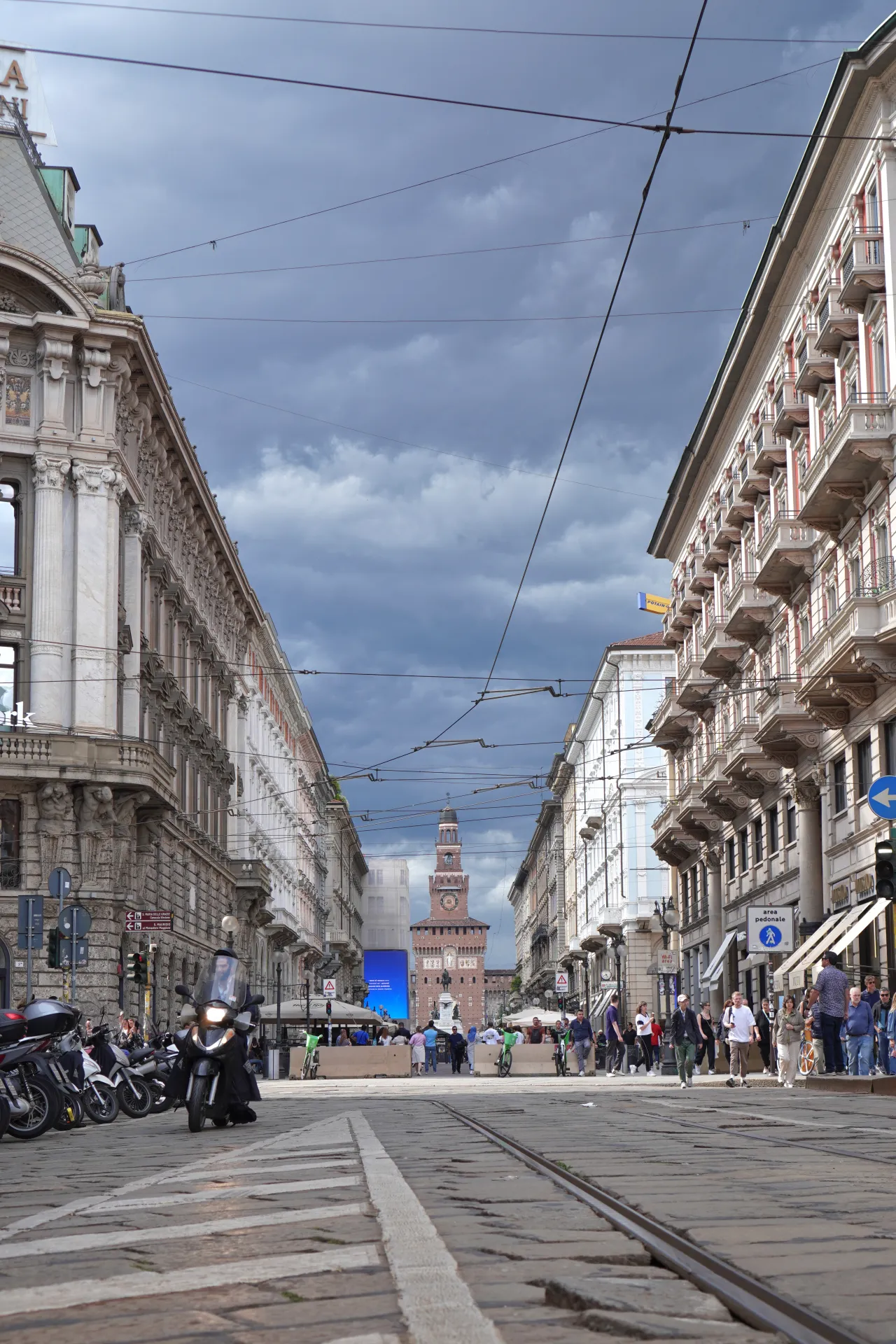 A street view of Via Dante in Milan, Italy, with Sforza Castle visible in the distance at the end of the avenue, flanked by ornate buildings and tram tracks on the cobblestone street.