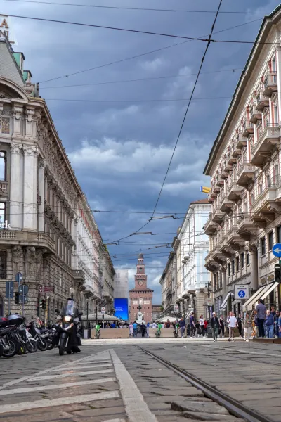 Una calle en Milán, Italia, con un tranvía que circula sobre vías de adoquines, flanqueada por edificios históricos. El Castillo Sforza es visible al fondo bajo un cielo nublado.