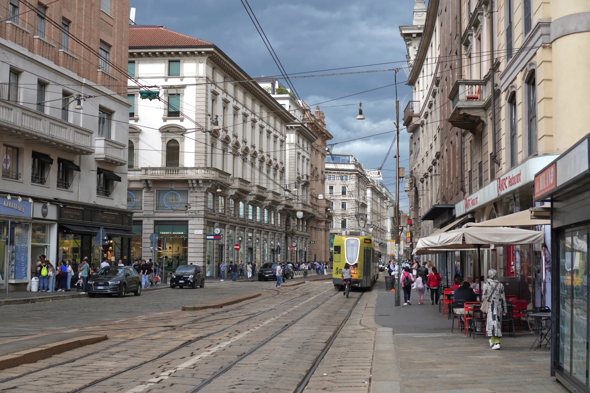 Ulica u Milanu, Italija, sa tramvajskim šinama koje prolaze sredinom. Zgrade se nižu sa obe strane ulice, u kojima se nalaze prodavnice i kafići. Ljudi hodaju trotoarima, a žuti tramvaj se kreće po šinama. Nebo je oblačno.