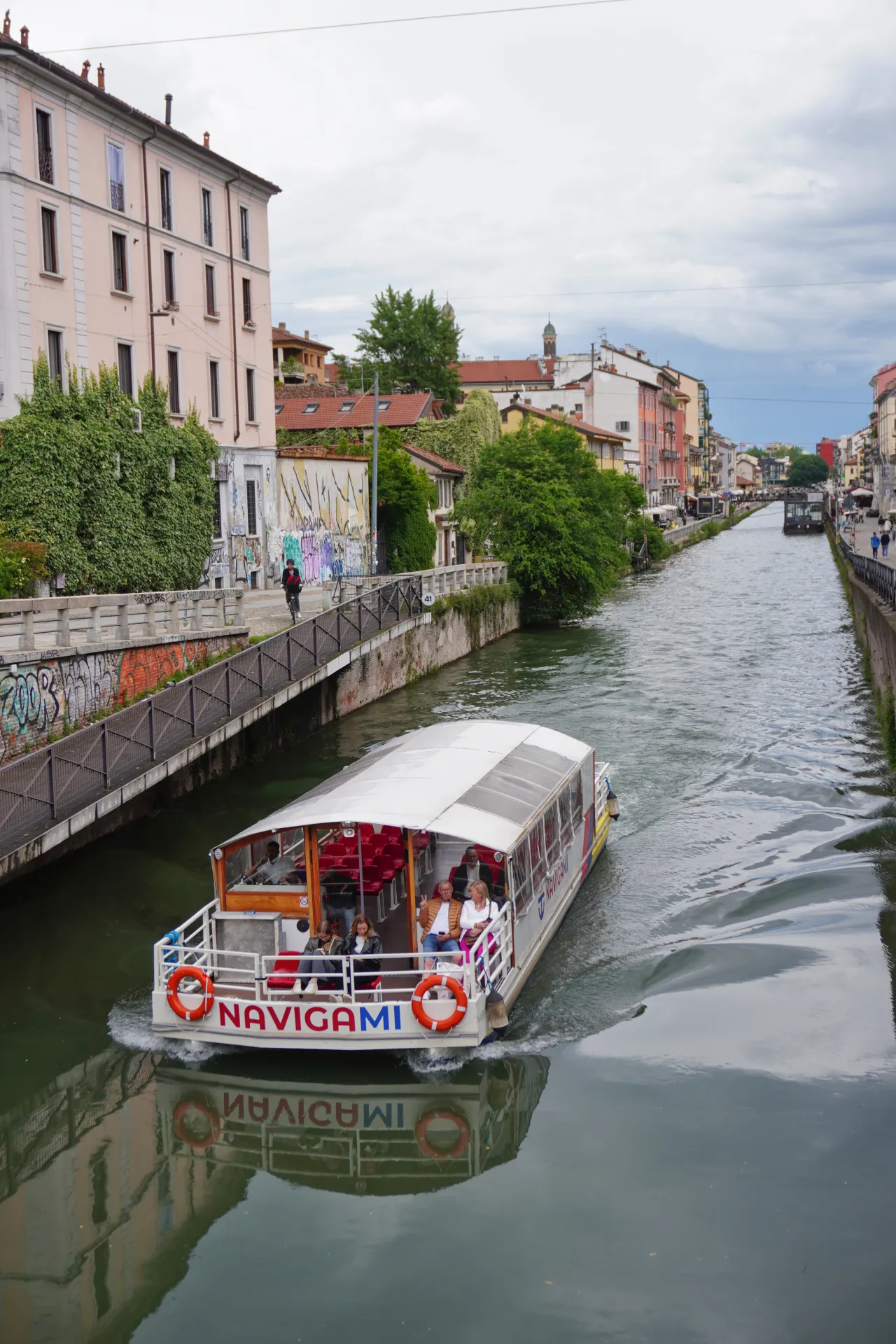A tour boat named "Navigami" travels on a canal in Milan, Italy, with passengers onboard. Buildings and greenery line the canal, and the boat's reflection is seen in the water.