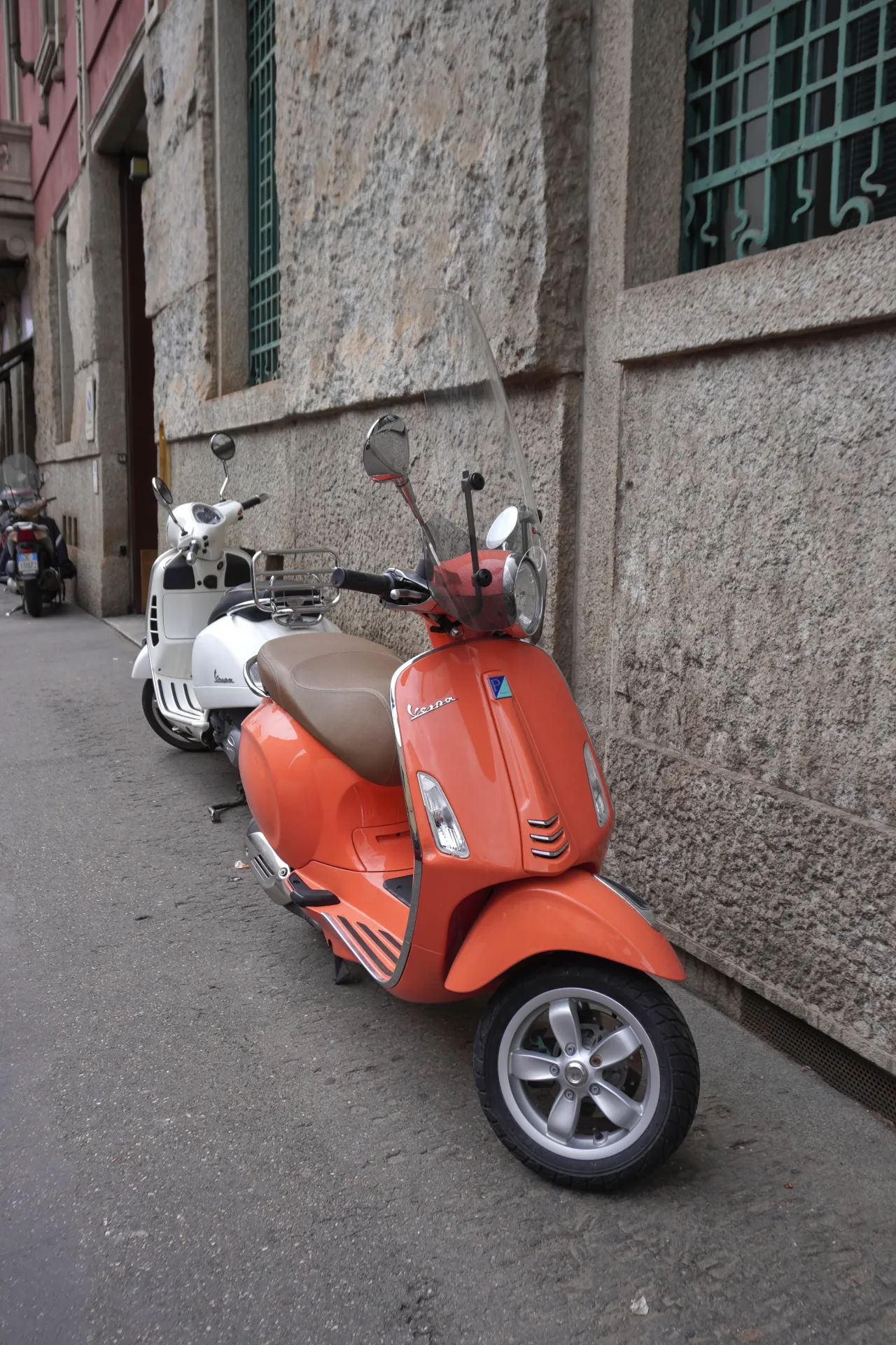 Narandžasti Vespa skuter u prvom planu i beli Vespa skuter iza njega, parkirani na ulici u Milanu, Italija. Vidljiva je kamena zgrada sa zelenom rešetkom na prozoru.