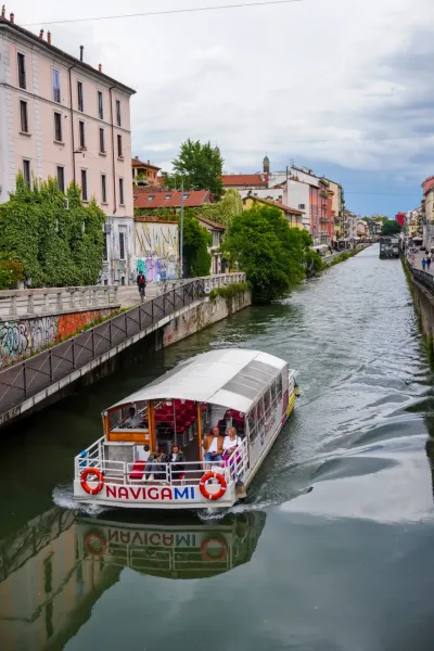 Un barco turístico llamado "Navigami" navega por un canal en Milán, Italia, con pasajeros a bordo. Edificios y zonas verdes bordean el canal, y el reflejo del barco es visible en el agua.