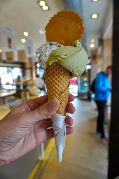 Una mano sostiene un cono de helado con tres bolas de gelato, incluyendo pistacho y crema de galleta, adornado con un barquillo redondo. En el fondo borroso, se ve a gente dentro de una heladería en Milán.
