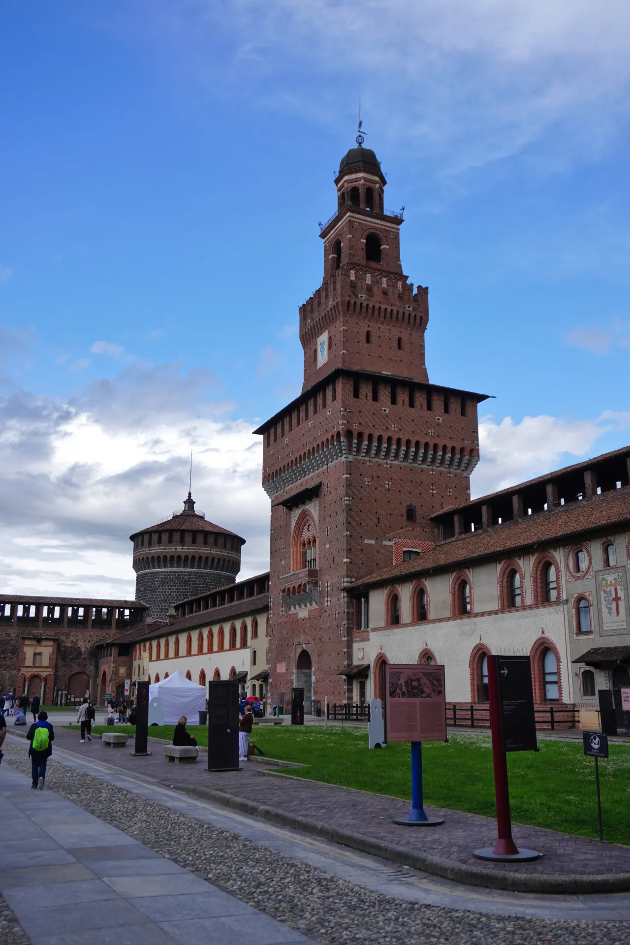 Sforza Castle with its tall brick tower and surrounding walls, a cobblestone path in the foreground and green grass with some informational signs.