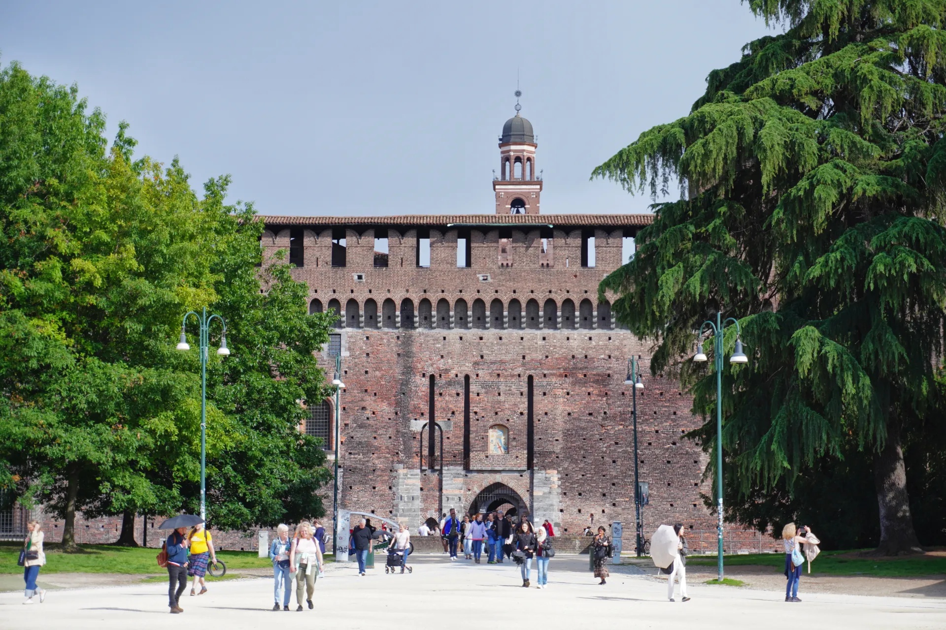 People walking in front of the Sforza Castle in Milan, Italy, with large trees framing the view.